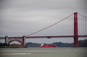 Golden Gate Bridge with red ship passing underneath on a cloudy day