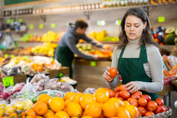 Young woman seller in uniform lays out tomatoes on counter in grocery store