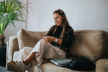 A young student prepares for school, sitting comfortably on a sofa. She is relaxed, wearing casual...