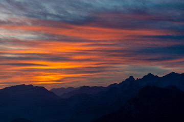 Sunset in the mountains. Red and orange burning sky horizon behind the alpine mountain range. Rocky mountain peaks of the Dolomites at sunset and dawn at sunrise light sunrays going through the clouds
