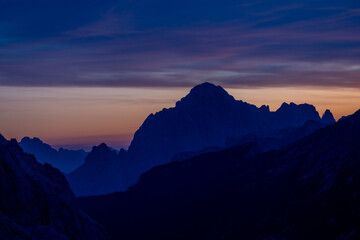 Sunset in the mountains. Red and orange burning sky horizon behind the alpine mountain range. Rocky mountain peaks of the Dolomites at sunset and dawn at sunrise light sunrays going through the clouds