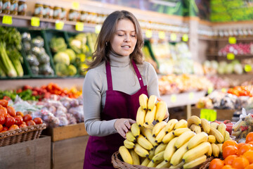 Young woman seller in uniform lays out bananas on counter in grocery store..