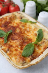 Delicious cooked lasagna in baking dish and basil on light grey table, closeup