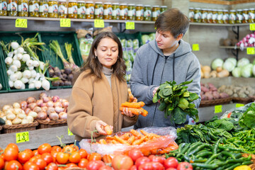 Happy young couple buying carrots and spinach in large fruit and vegetable market