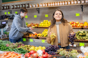 Young woman shopper in casual clothes chooses grapes in vegetable shop