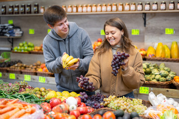 Couple of young woman and young guy choose grapes and bananas in vegetable shop..