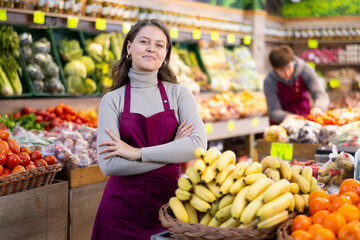 Happy young female seller standing by basket full of bananas in large grocery market