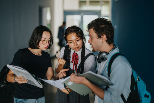Three diverse students engage in an animated discussion, holding notebooks and textbooks, while standing in a busy school hallway. The scene captures academic collaboration and lively student