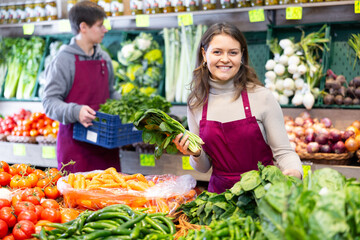 Smiling female seller in uniform working in grocery shop, posing with salad and spinach