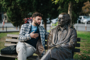 A man engages in a lively discussion with a bronze statue on a park bench, symbolizing contemplation and imagination in an urban setting.