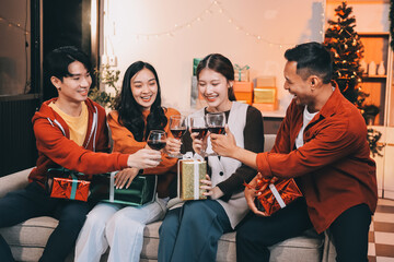 Group of young Asian man and women as friends having fun at a New Year's celebration, holding gift boxes standing by Christmas tree decoration, midnight countdown Party at home with holiday season.