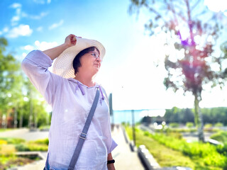 A woman is playfully adjusting her large sun hat while standing in a vibrant park filled with trees...