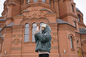 A woman stands outdoors, capturing a photo of a large red brick building while dressed in warm clothing amid a cool atmosphere