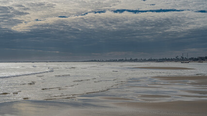 The low tide in the ocean. The waves roll towards the shore, spreading out. Rivulets and puddles of water form a pattern on the sand. Silhouettes of people and boats in the distance. Blue sky, clouds.