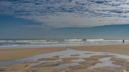 The low tide in the ocean. Puddles of water on a sandy beach. A fishing boat is visible among the foaming waves. A man on the shore pulls out a fishing net. Blue sky, clouds. Madagascar. Morondava. 