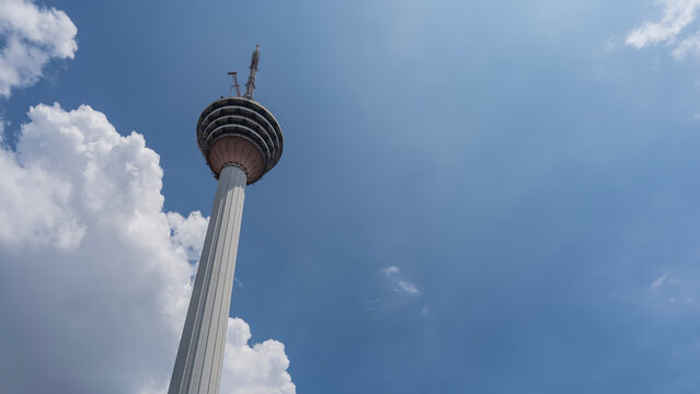 A tall TV tower on a background of blue sky and clouds. The spire and the restaurant is on top. Menara Kuala Lumpur. Bottom-up view.  Malaysia