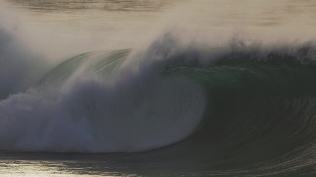Large wave barrels forward as mist trails off the crest, surfers visible in lineup