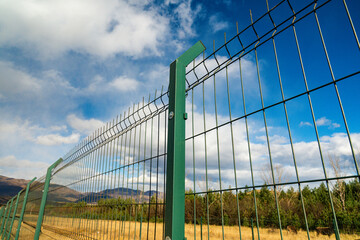 Fototapeta premium A close-up view of a green metal fence against a vivid sky and mountains, highlighting security and separation concepts