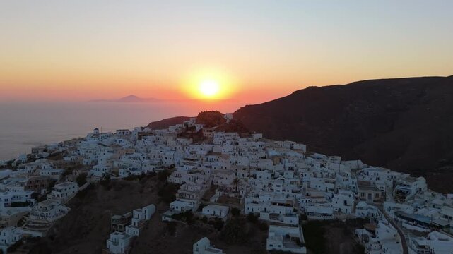 Aerial establishing overview of Anafi coastline at sunset with sun casting warm glow over rugged terrain with iconic white homes, Greek Island scenery