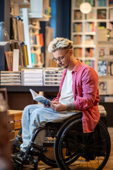 Focused young man with disability reading book in bookstore. Interested student guy in wheelchair...