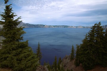 View of massive crystal clear blue lake from high elevation, showing pine trees on the nearby ridge, a dramatic cloudscape over head and in the far distance the other side of the volcanic crater. 