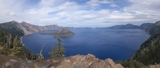 Panoramic view of beautiful crystal clear blue lake from high elevation, showing pine trees on the nearby ridge, a dramatic cloudscape over head and in the far distance the other side of the volcanic 