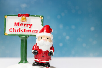 Miniature Santa Claus figurine standing next to a festive Merry Christmas sign with a snowflake-filled blue background, perfect for holiday-themed designs or decorations.