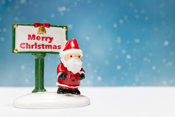 Miniature Santa Claus figurine standing next to a festive Merry Christmas sign with a snowflake-filled blue background, perfect for holiday-themed designs or decorations.