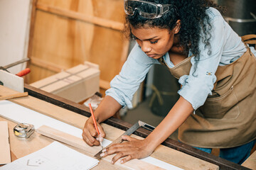 African American carpenter woman drawing a line on wood with a pencil and ruler in workshop. Focused on precise measurements, woodworking, and construction tasks in carpentry, National Carpenters Day