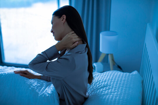 Young woman experiencing neck pain, sitting on bed at night, suffer aching and tightness in shoulders, having muscle spasms and discomfort