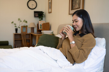 Pleased young woman enjoying warm drink sitting under a white duvet in bright bedroom