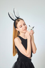 Obraz premium A young beautiful girl in a bunny costume and black clothes on a clean white background. In the hands of a small rabbit