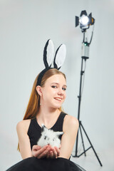A young beautiful girl in a bunny costume and black clothes on a clean white background. In the hands of a small rabbit
