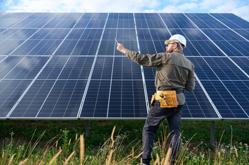 Engineer in uniform with protective helmet near solar station holding screwdriver. Supervisor is checking on solar panels on solar farm. Green energy concept.