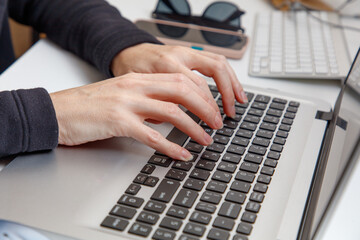 A person is typing on a laptop with a keyboard and a cell phone on the desk