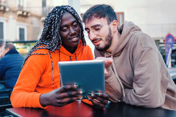 Young African woman and Caucasian man using digital tablet together outdoors sitting at urban cafe table