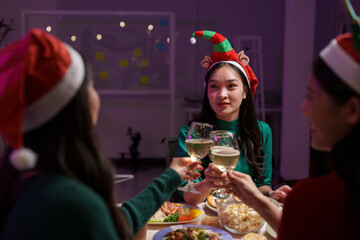 A group of Asian friends are celebrating the New Year, holiday, Christmas together. Young people wearing Santa hats happily clinking wine glasses at the dining room table in the living room.