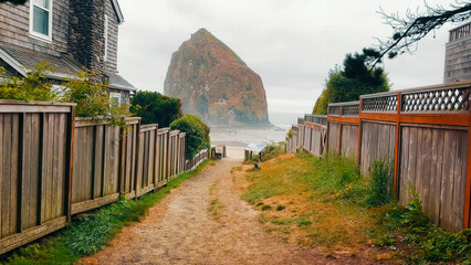 Pathway to Haystack Rock on a cloudy day. A picturesque pathway leads down to the beach, framed by wooden fences and coastal homes, with Haystack Rock towering in the distance.
