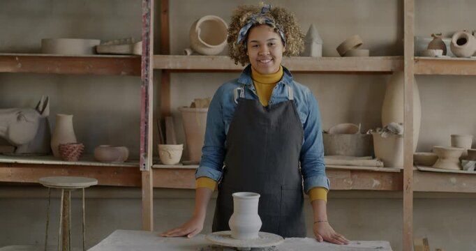 Slow motion portrait of cheerful African American woman in apron standing in pottery workshop next to throwing wheel smiling looking at camera