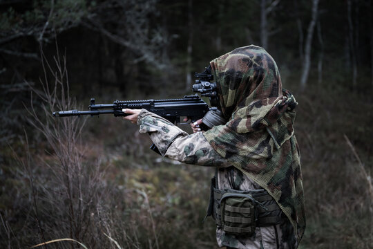 A woman soldier in a gas mask, camouflage and a tactical helmet with a night vision monocular in the forest at dusk.