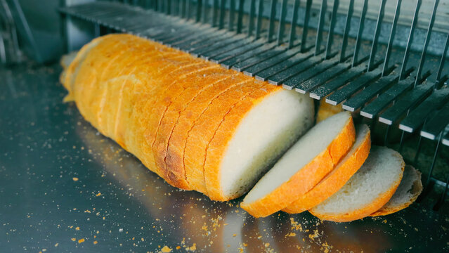 Freshly Sliced Bread Loaf in a Commercial Bread Slicer. A close-up of a loaf of crusty white bread being sliced evenly in a commercial bread slicer with crumbs scattered.