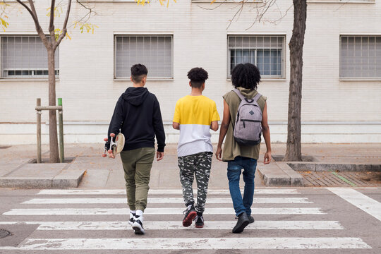 rear view of three diverse teen friends with backpack and skateboard crossing road on crosswalk, concept of youth and urban lifestyle
