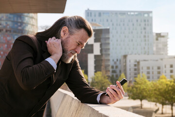 concerned mature male with beard in classy suit using mobile phone while standing against modern...
