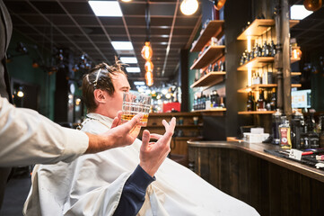 Exclusive offer of drinks from luxury barbershop. Young client taking a glass of whiskey from hairdresser. Side view of male customer getting new haircut.