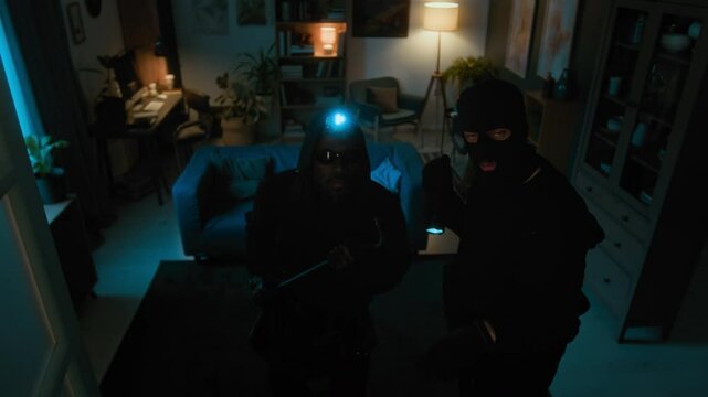 High angle view of two diverse male thieves moving around dark apartment, man in black balaclava noticing security camera, shining at it with flashlight and tapping finger on it