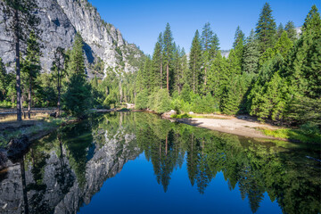 Serene view of the mirror like Merced River reflecting pine trees and granite cliffs in Yosemite National Park, California, on a bright summer morning