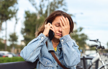 Urban woman sitting on a bench with her hand on her forehead, listening to bad news on her mobile phone with a distressed expression.