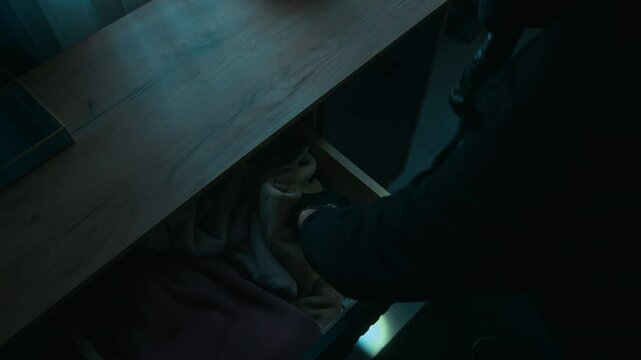 View from above of male thief rummaging through dresser drawer using flashlight, then finding two packs of money and leaning into camera with happy look