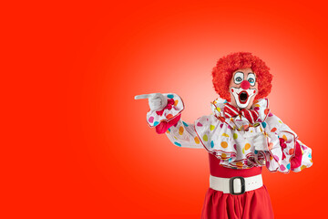 Surprised clown pointing at blank template copy spase for advertising, posters, greetings, invitations. Entertainer as Joker in a suit and wig, with clown whiteface makeup