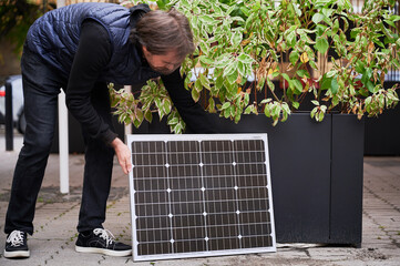 Man crouches down to place solar panel near large planter with green foliage. Guy in black outfit...